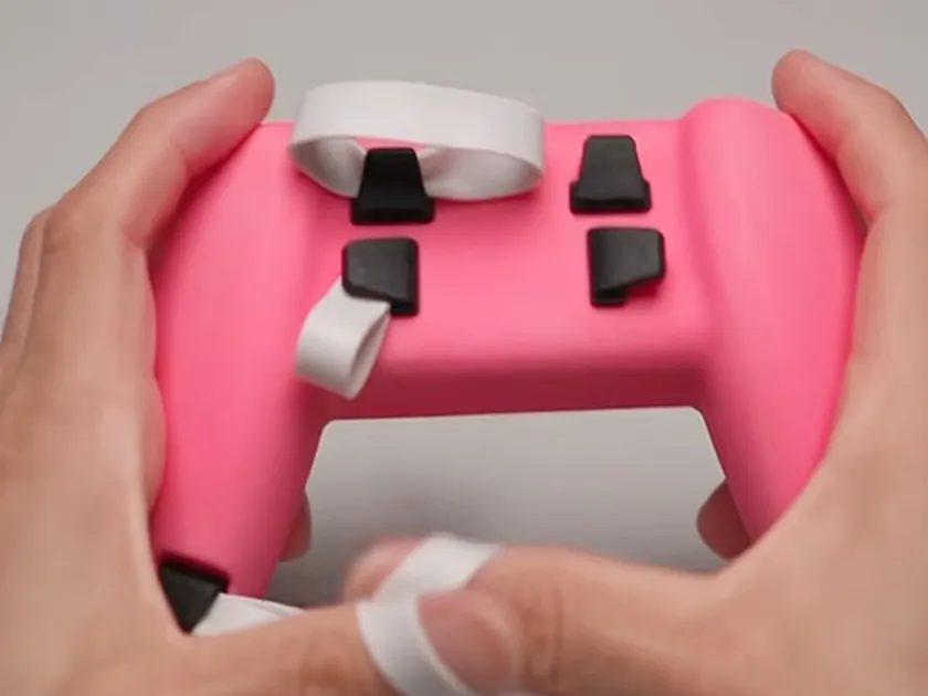 A 3D-printed G Hands prototype in pink, held in both hands, with black band hooks and white finger bands looped through them.